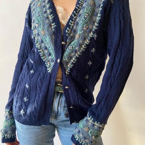 Könnte beinhalten: Marineblauer Cardigan mit aufwendiger Blumenstickerei in Blau-, Grün- und Beigetönen. Der Pullover hat ein Zopfmuster an Körper und Ärmeln mit Knopfverschlüssen. Die Manschetten und der Halsausschnitt sind mit detaillierter Stickerei verziert.