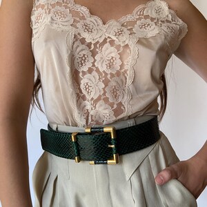 May include: A beige lace camisole with a green snakeskin belt. The belt has a gold buckle.