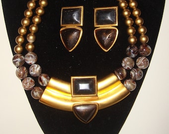 May include: A vintage jewelry set featuring a gold-tone necklace and matching earrings. The necklace has two strands of gold beads and a gold-tone pendant with a dark square and triangle accent. The earrings have a square top and a triangle bottom.