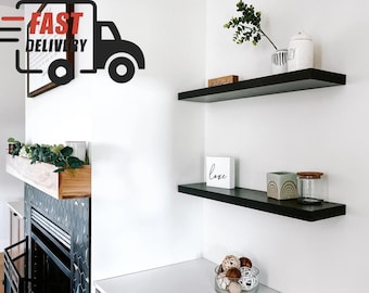 May include: Two black floating shelves mounted on a white wall. The top shelf has a white ceramic jar with a black and white design, a small wooden sign that says "blessed", and a small white ceramic jar. The bottom shelf has a white sign that says "love", a green and brown ceramic jar with a rainbow design, and a clear glass jar with a brown lid.