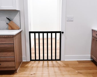 May include: A black metal baby gate with vertical bars is installed in a doorway between a kitchen and a hallway. The gate is open and the hallway is visible through the gate.