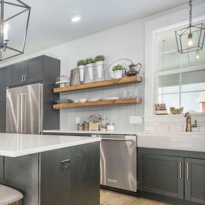May include: A modern kitchen with a large island, wooden chairs, and pendant lights. The island has a white countertop and dark gray cabinets. The kitchen features stainless steel appliances, wooden shelves, and a farmhouse sink. The flooring is light-colored wood.