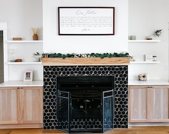 Op de afbeelding: Een woonkamer met een zwarte zeshoekige tegelomranding en een houten mantel. Boven de mantel is een ingelijst bord met tekst. De kamer beschikt over ingebouwde kasten en zwevende planken.