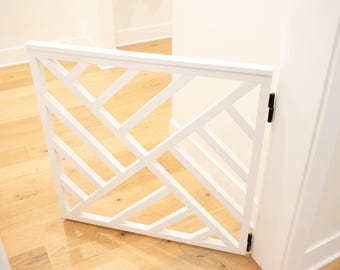 May include: A white wooden baby gate with a geometric design. The gate is open, revealing a light wood floor and white walls. The gate is attached to a white door frame with black hinges.