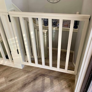 Op de afbeelding: Witte houten babyhek met verticale latten, ge&iuml;nstalleerd bovenaan een met tapijt beklede trap. Het hek is gesloten en heeft een witte rand rond de boven- en onderkant.