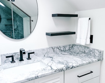 May include: A white bathroom vanity with black hardware and a white marble countertop. The vanity has two drawers and a cabinet with louvered doors. There is a black faucet and a white porcelain sink. The vanity is in front of a white wall with two black floating shelves and a black towel rack with a white towel.