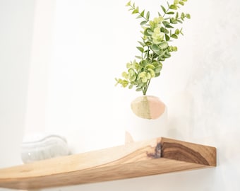 Op de afbeelding: Een houten plank met een lichtroze en groene keramische vaas met groene bladeren. De plank is gemonteerd aan een witte muur.