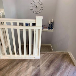 Op de afbeelding: Een witte houten babyhek met een witte leuning en een witte paal op een houten vloer. Het hek staat voor een met tapijt beklede trap. Er is een plank met kaarsen aan de muur rechts van het hek.