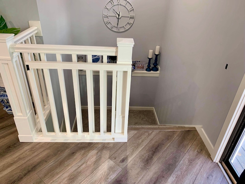 Op de afbeelding: Een witte houten babyhek met een witte leuning en een witte paal op een houten vloer. Het hek staat voor een met tapijt beklede trap. Er is een plank met kaarsen aan de muur rechts van het hek.