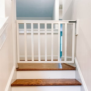 May include: White wooden baby gate with a black metal latch installed at the top of a wooden staircase.