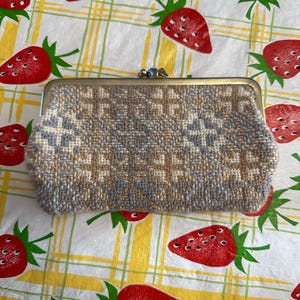 May include: A small, gray and beige woven clutch purse with a metal clasp. The purse is on a white tablecloth with a red strawberry pattern.
