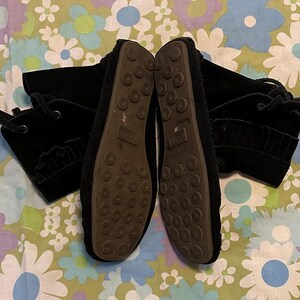 May include: A pair of black suede ankle boots with laces and tassels. The boots have a textured sole and are shown on a floral patterned background.