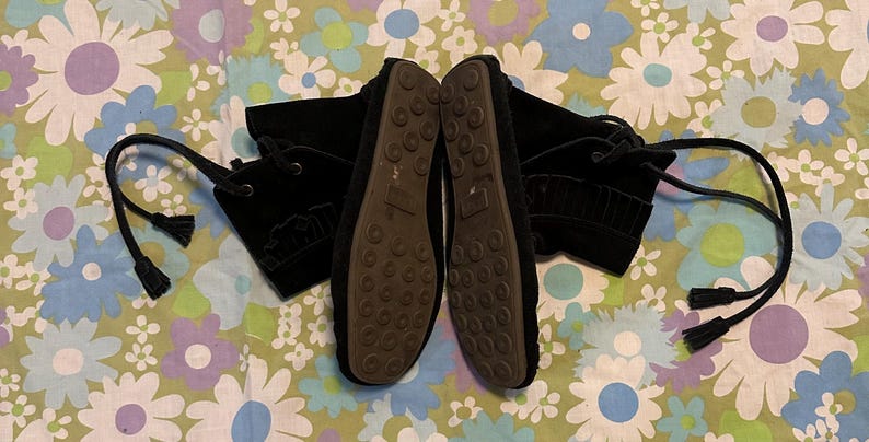 May include: A pair of black suede ankle boots with laces and tassels. The boots have a textured sole and are shown on a floral patterned background.