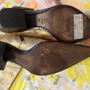Puede incluir: Botines dorados con tac&oacute;n de bloque. Los botines tienen punta afilada y cremallera lateral. Las suelas son marrones y el tac&oacute;n mide unos 5 cm de alto.