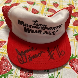 May include: A red and white trucker hat with the text "Team Motorsports Wear It!" in black. The hat is signed "Jamie Swanson #16" in black marker.