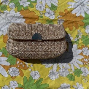 May include: A beige woven clutch with a silver-toned clasp. The rectangular bag has a flap closure and a textured, grid-like pattern. The clutch is set against a yellow floral background.