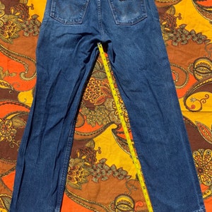 Puede incluir: Un par de pantalones vaqueros de mezclilla azul oscuro con una cinta m&eacute;trica amarilla colocada sobre la parte posterior de los pantalones. Los pantalones tienen un parche de cuero marr&oacute;n con la palabra "Wrangler" en el bolsillo trasero derecho.