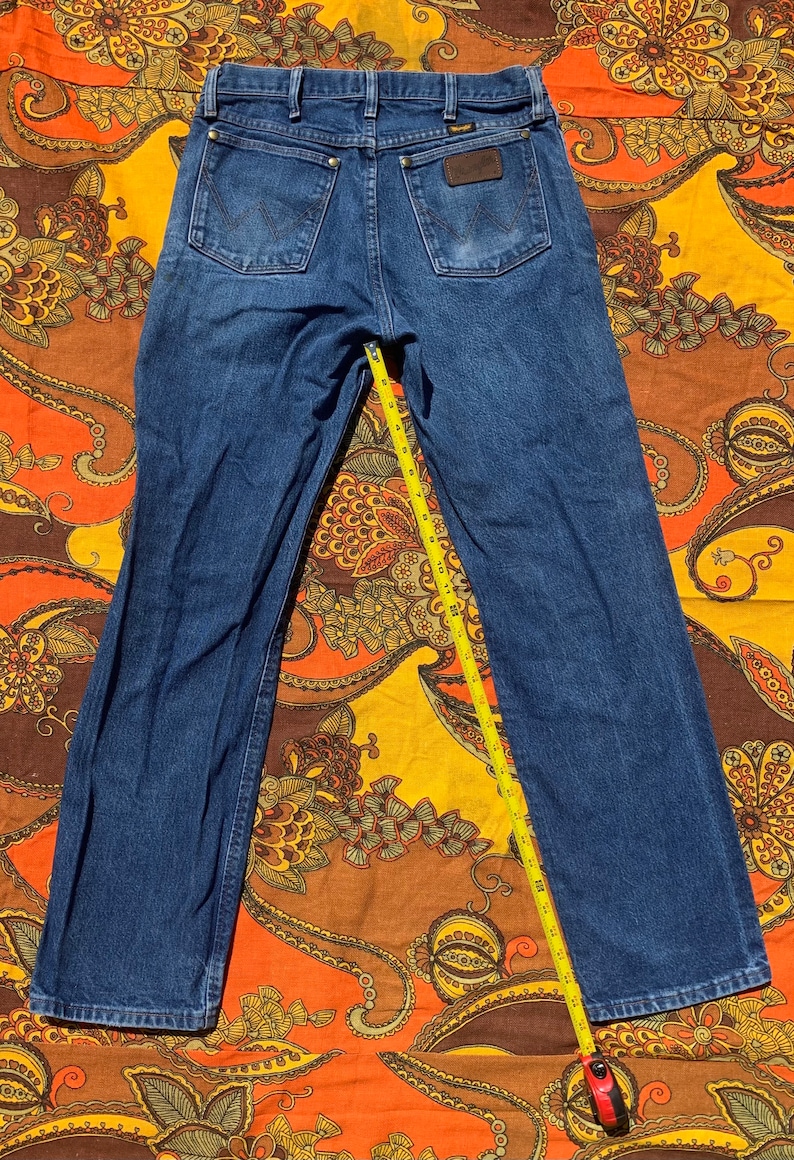 Puede incluir: Un par de pantalones vaqueros de mezclilla azul oscuro con una cinta m&eacute;trica amarilla colocada sobre la parte posterior de los pantalones. Los pantalones tienen un parche de cuero marr&oacute;n con la palabra "Wrangler" en el bolsillo trasero derecho.