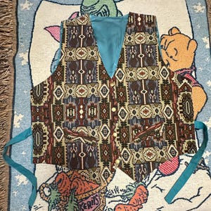 Könnte beinhalten: Eine ärmellose Weste mit einem mehrfarbigen geometrischen Webmuster in Braun-, Blau- und Cremetönen. Das Kleidungsstück hat ein einfarbiges türkisblaues Innenfutter, einen V-Ausschnitt, fünf goldfarbene Knöpfe vorne und zwei seitliche Bindebänder.
