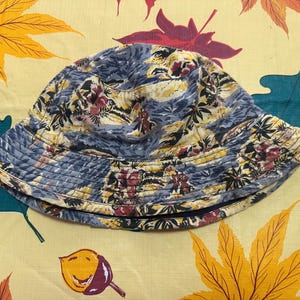 Peut inclure: Un chapeau bob avec un imprimé tropical multicolore représentant des palmiers, des fleurs et du feuillage dans des tons de bleu, jaune et rouge. Le chapeau présente une calotte arrondie et un bord incliné avec des coutures horizontales visibles.