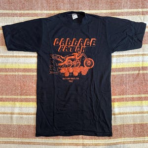 May include: Black t-shirt with an orange graphic featuring the text "Cabbage Patch" above a motorcycle design. Below the design is "Daytona Beach, Fla. 1982". The shirt has short sleeves and a crew neck.