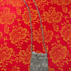 May include: A silver beaded crossbody bag with a drawstring closure and a long, thin strap. The bag has two small, round, decorative beads near the bottom. The background is a red fabric with a floral pattern.