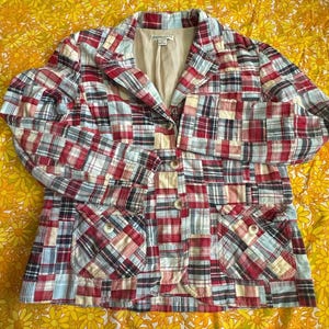 May include: A patchwork blazer in red, white, blue, and black plaid patterns. The jacket has a notched collar, two front pockets, and three button closures. The lining is a solid beige color.