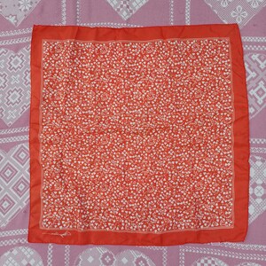 May include: A square scarf in a vibrant orange-red hue featuring a dense, repeating white pattern of small heart-shaped leaves and delicate vines. The design is framed by a thin white border, and a small signature is visible in the corner.