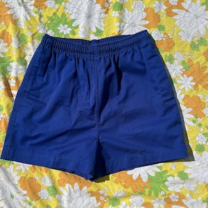 May include: Royal blue elastic waist shorts. The shorts are made of a solid, smooth fabric. The waistband is gathered with elastic. The shorts are laid flat on a floral patterned sheet with white, orange, and green flowers.