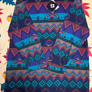 Peut inclure: Un t-shirt à manches longues et col rond arborant un motif géométrique répétitif dans des tons bleu marine, sarcelle, magenta et beige. Le vêtement comporte une étiquette noire avec la lettre M blanche et une étiquette de col avec les lettres BE. Le design se compose de bandes horizontales de zigzags, de losanges et de formes stylisées.
