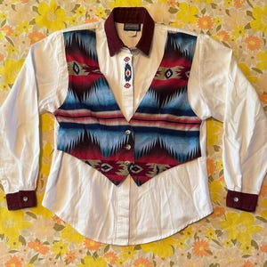 May include: White button-down shirt with a burgundy collar and cuffs. The shirt features a colorful vest with a geometric pattern in red, blue, and black. The vest has silver buttons.