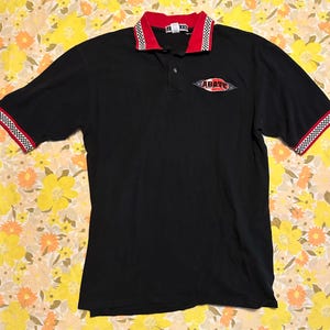 May include: Black polo shirt with red and checkered accents on the collar and sleeves. The shirt features the word "ABATE" in a stylized design on the chest.