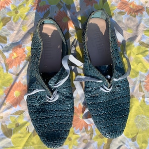 May include: A pair of dark blue woven leather shoes with white laces. The shoes have a unique, textured design and are sitting on a floral patterned fabric.