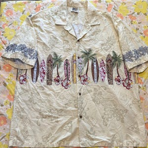 Könnte beinhalten: Beige Hemd mit Knöpfen und tropischem Design. Das Hemd hat ein Muster aus Surfbrettern, Palmen und Hibiskusblüten. Die Ärmel haben eine Blumenborte. Das Hemd hat eine lockere Passform und einen klassischen Kragen.