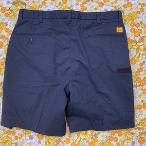 May include: Navy blue work shorts with a button closure, belt loops, and back pockets. A small black pocket is on the right side. The shorts have a tag that reads "Dickies".