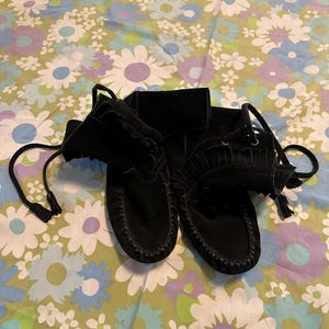 May include: A pair of black suede moccasins with fringe and laces. The moccasins are on a floral patterned fabric.