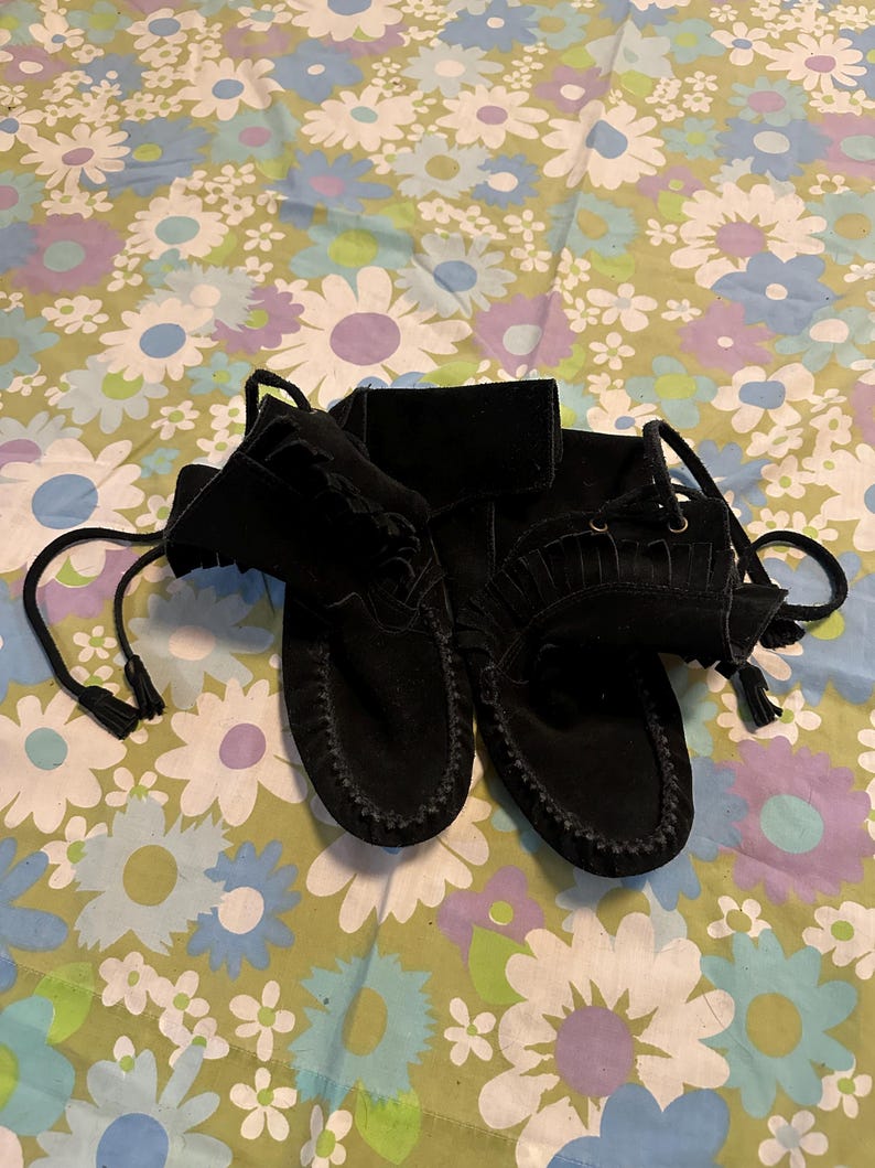 May include: A pair of black suede moccasins with fringe and laces. The moccasins are on a floral patterned fabric.