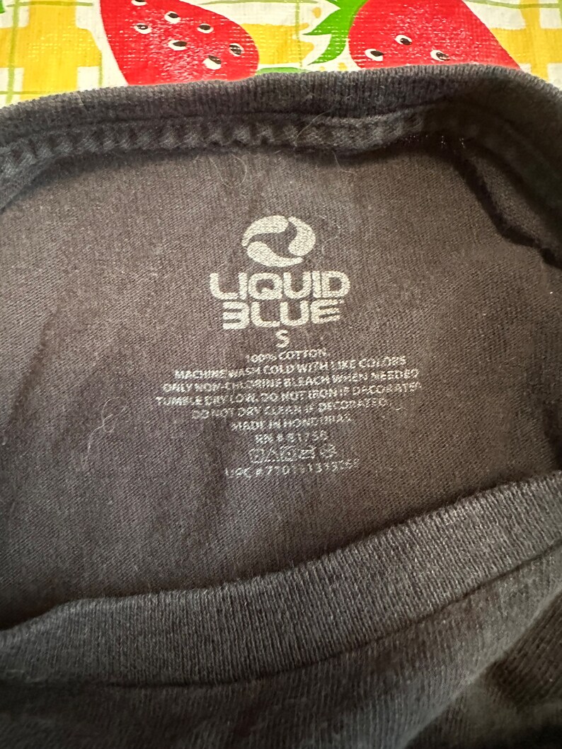 Puede incluir: Una etiqueta de camiseta negra Liquid Blue con texto blanco. La etiqueta dice "Liquid Blue", "S", "100% Algod&oacute;n", "Lavar a m&aacute;quina con agua fr&iacute;a con colores similares", "Usar solo blanqueador sin cloro cuando sea necesario", "Secar en secadora a baja temperatura. No planchar si est&aacute; decorado", "No lavar en seco si est&aacute; decorado", "Hecho en Honduras", "RN#1756", y "UPC #770151339758".