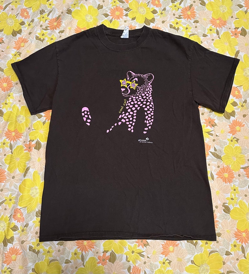 Pu&ograve; includere: Una maglietta marrone con un ghepardo rosa che indossa occhiali da sole e la scritta "cool cat" stampata sul davanti. La maglietta ha un piccolo logo che dice "get outside nature" nell'angolo in basso a destra.