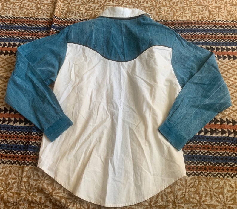 May include: A long-sleeved shirt with a white body and blue sleeves and yoke. The shirt has a button-down collar and a curved yoke design. The sleeves and yoke are a textured blue fabric, while the body is a smooth white fabric.