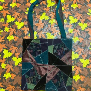 May include: A patchwork tote bag with a variety of fabrics in shades of green, brown, and black. The bag has long straps in teal and brown. The background is a brown surface with yellow and orange leaf patterns.