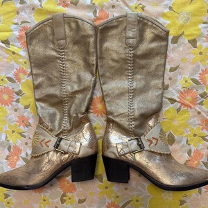 Puede incluir: Un par de botas de vaquero doradas hasta la rodilla. Las botas tienen un acabado met&aacute;lico texturizado, costuras decorativas y una hebilla. Los tacones miden aproximadamente 7,6 cm de alto.