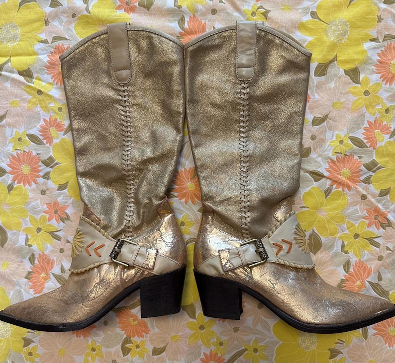 Puede incluir: Un par de botas de vaquero doradas hasta la rodilla. Las botas tienen un acabado met&aacute;lico texturizado, costuras decorativas y una hebilla. Los tacones miden aproximadamente 7,6 cm de alto.