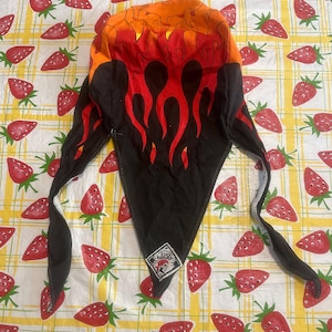 Pode incluir: Um bandana preto com chamas laranja e vermelhas impressas nele. O bandana tem uma etiqueta branca com o texto "Genuine Do-Wrap" e uma imagem de desenho animado de uma pessoa usando um bandana.