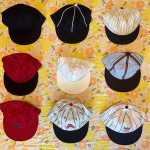 May include: Nine baseball caps in various colors and designs are displayed. The caps include solid black, white-lined black, pinstriped, red with black brim, white, gray with red trim, red with green emblem, striped with an "A", and striped with a shield.