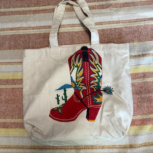 May include: Off-white canvas tote bag featuring a red cowboy boot design with yellow and green accents, a spur, and a cactus. The bag has two handles and is suitable for carrying groceries or personal items.