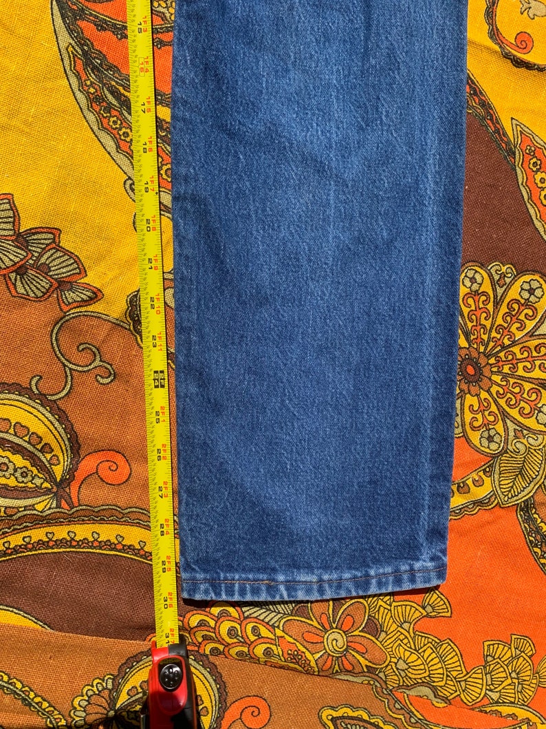 Puede incluir: Primer plano de la parte inferior de un par de pantalones vaqueros azules. Se puede ver una cinta m&eacute;trica en el lado izquierdo de la imagen.