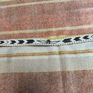 May include: A beaded belt with a white background and black arrow and dragonfly designs. The belt is displayed on a striped fabric with orange, yellow, and cream colors.