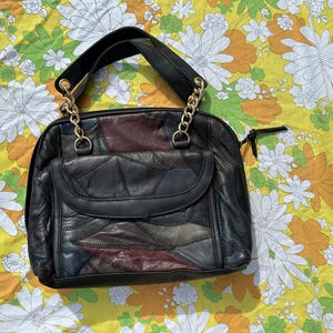 Könnte beinhalten: Schwarze Lederhandtasche mit einem Patchwork-Design in verschiedenen Farben. Die Tasche hat einen oberen Griff und einen goldfarbenen Kettenriemen. Der Hintergrund ist ein gelbes Blumenmuster.