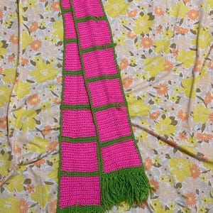 Peut inclure: Une écharpe en crochet rose et verte avec des franges. L'écharpe est faite de carrés avec une bordure verte.
